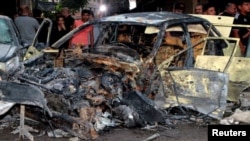 Orang-orang memperhatikan kendaraan yang hangus terbakar di lokasi ledakan bom di luar Masjid Sayeda Zeinab di selatan Kota Damaskus, Ibu Kota Suriah, 27 Juli 2023. (Foto: SANA/handout via Reuters)