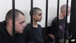 Двоє громадян Великобританії Ейден Аслін (ліворуч) і Шон Піннер (праворуч) і марокканець Саудун Брагім (в центрі) сидять за ґратами в залі суду в Донецьку, на території, що підконтрольна уряду ДНР, на сході України, четвер, 9 червня 2022 р. (AP Photo)